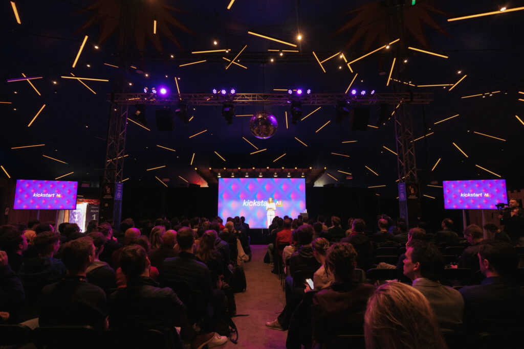 Een publiek van mensen kijkt naar een spreker op het podium tijdens het 'Kickstart AI' evenement. De zaal is verlicht met paarse en gele lichten die een sterrenhemel-effect creëren