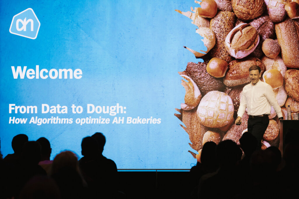 Simon Grest from Albert Heijn stands on stage next to a large screen displaying the text 'From Data to Dough: How Algorithms optimize AH Bakeries' with an image of various breads.