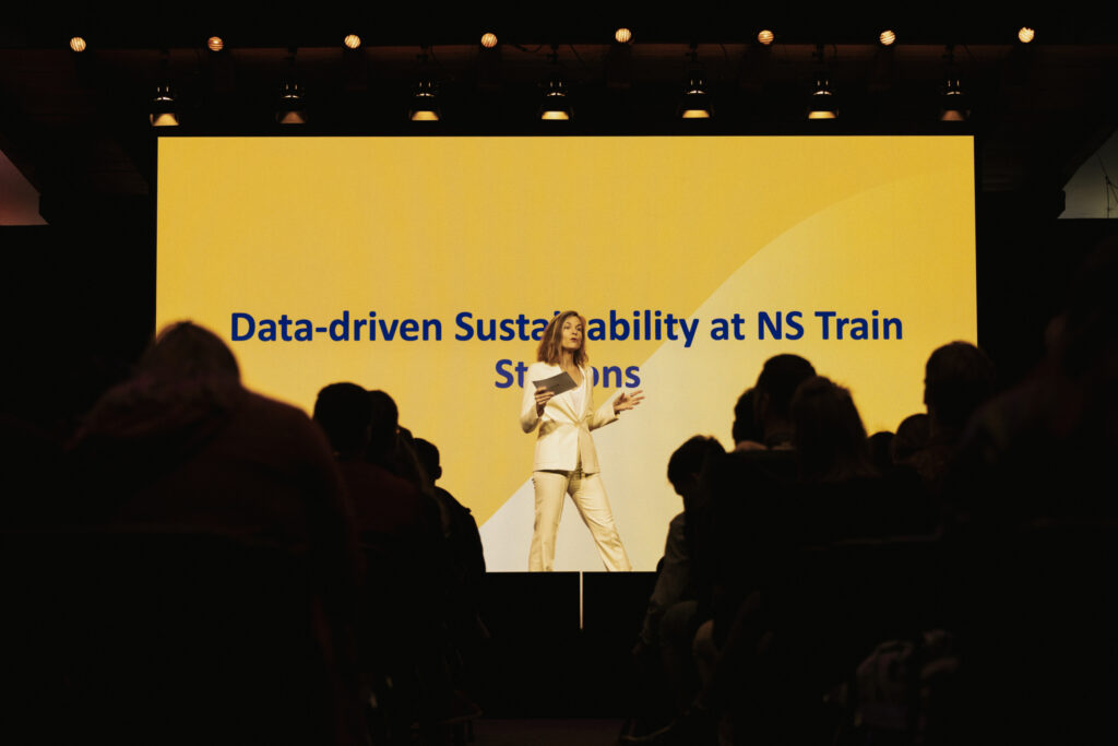 Irene Rompa stands on stage in front of a large screen with the text 'Data-driven Sustainability at NS Train Stations,' introducing Branka Milivojevic, data scientist at NS.
