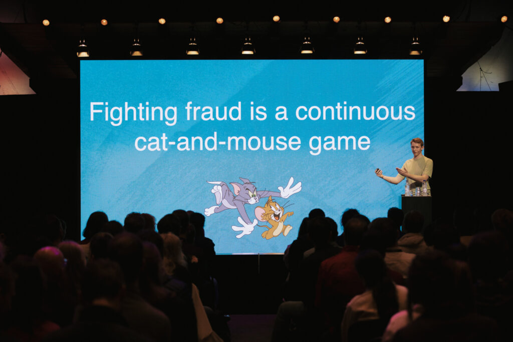 A speaker, Martynas Jašinskas, data scientist at Vinted, stands on stage in front of a large screen displaying the text 'Fighting fraud is a continuous cat-and-mouse game' with an image of Tom and Jerry.
